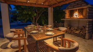 An outdoor dining area with a wooden table set for four, surrounded by cushioned chairs, and a stone fireplace with logs.