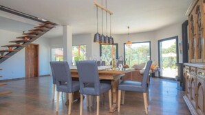 A spacious dining area with a wooden table and chairs, a staircase, and large windows offering natural light.