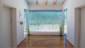 A hallway with wooden flooring, a ceiling with exposed beams, and large windows with horizontal blinds.