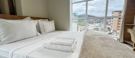 A hotel room with a large bed, a view of the city through a large window, and a wooden headboard.