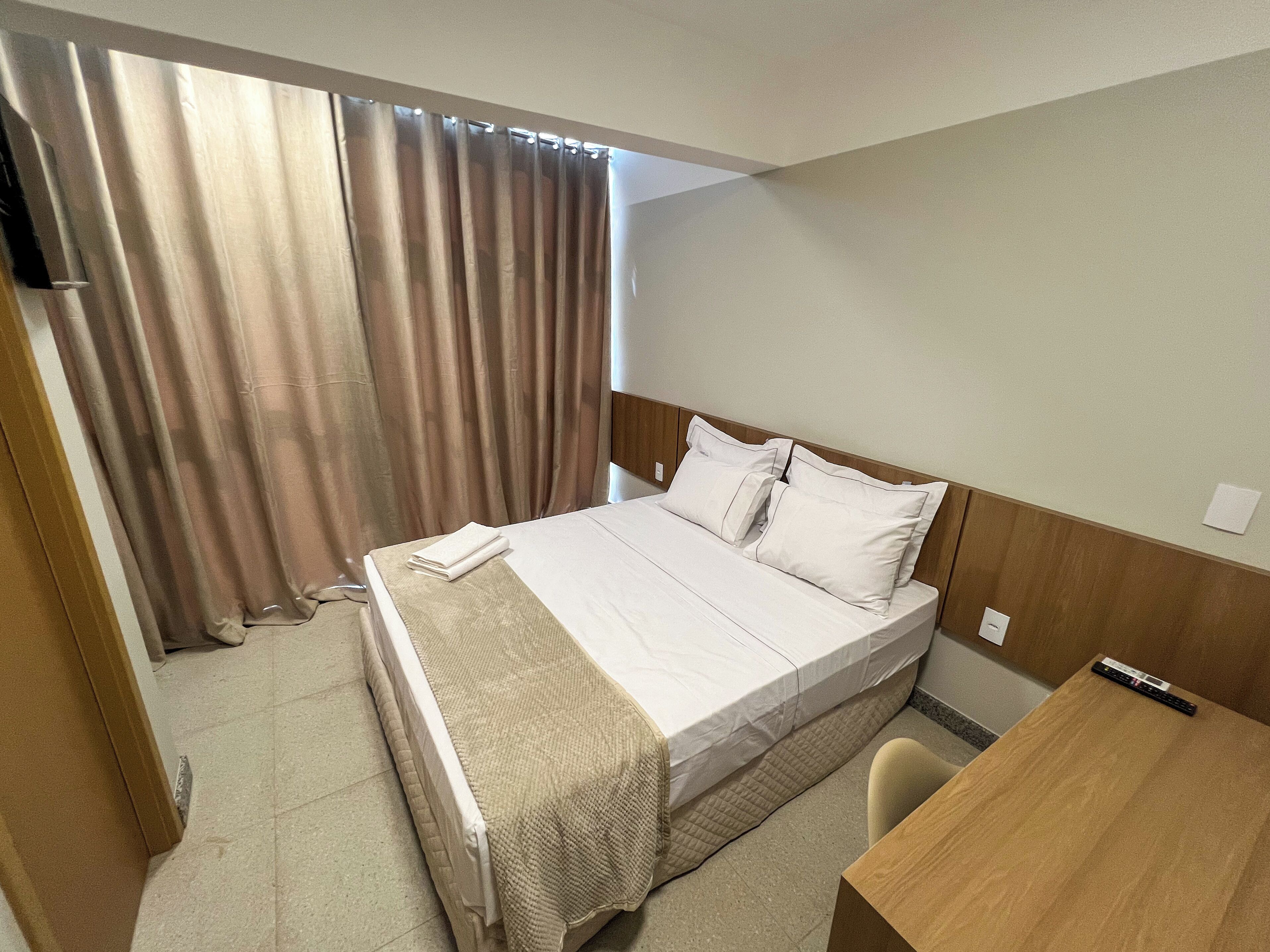 A neatly made bed with white linens and pillows, a wooden headboard, a small wooden desk, and a curtain partially drawn to the side.