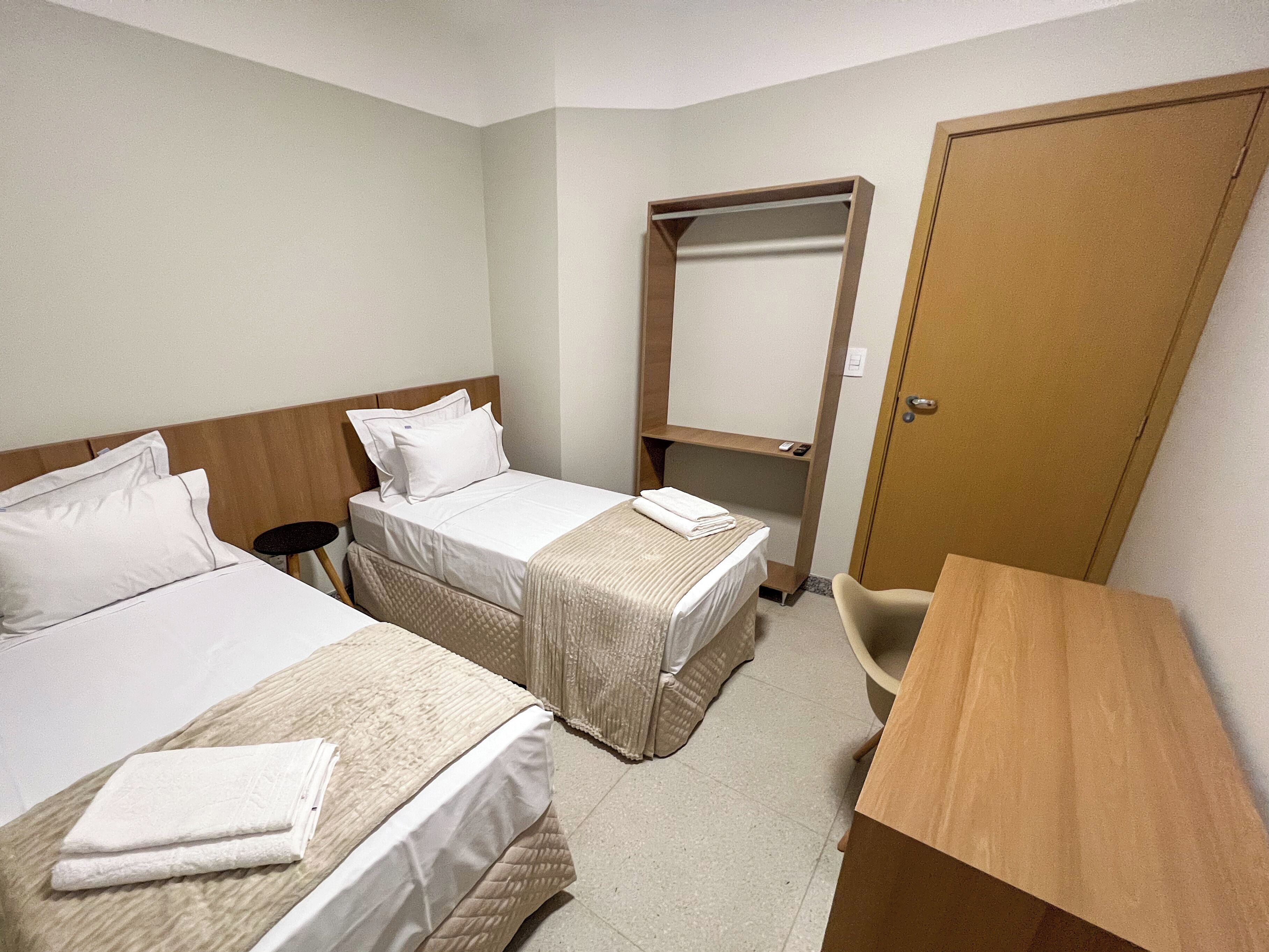 Quarto de hotel com duas camas, uma escrivaninha, uma cadeira, um guarda-roupa e uma mesinha.
