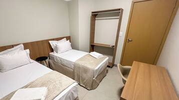 Quarto de hotel com duas camas, uma escrivaninha, uma cadeira, um guarda-roupa e uma mesinha.