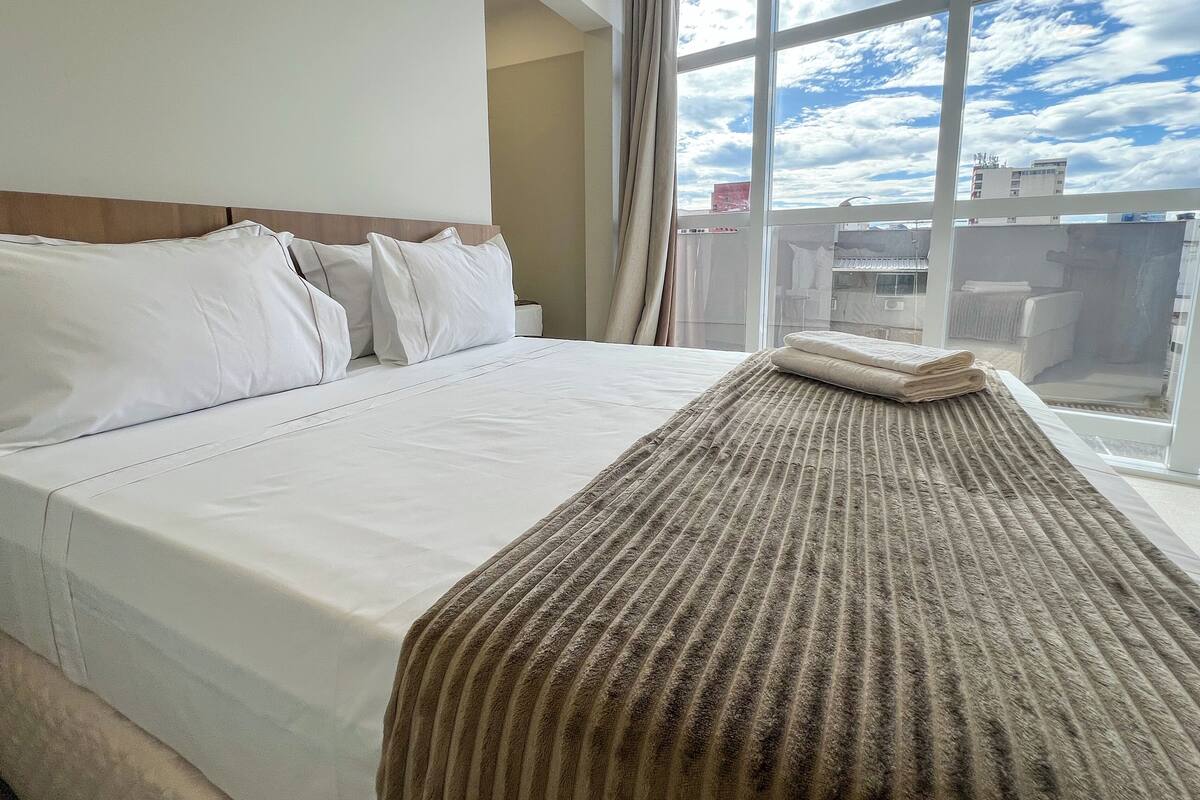 A hotel room with a large bed, a view of the sky through a window, and a balcony with a railing.