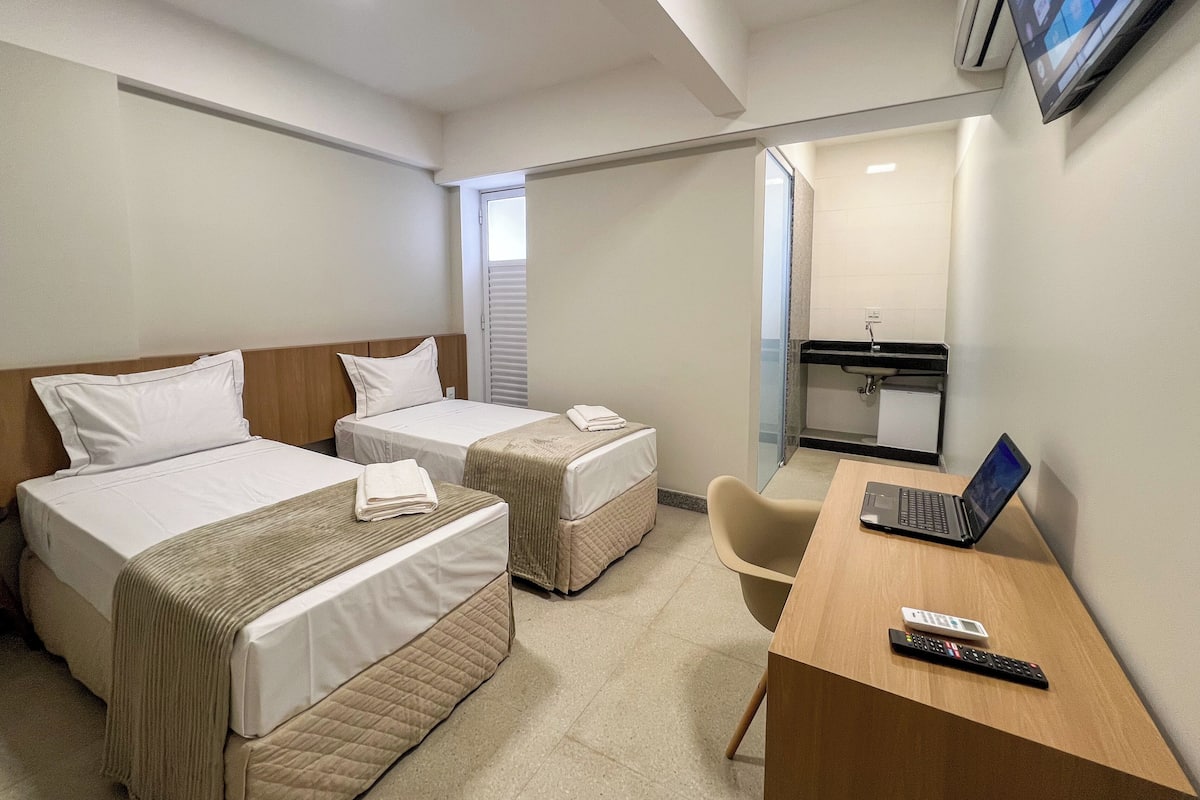 A hotel room with two beds, a desk with a laptop, a TV, and a bathroom with a sink and mirror.
