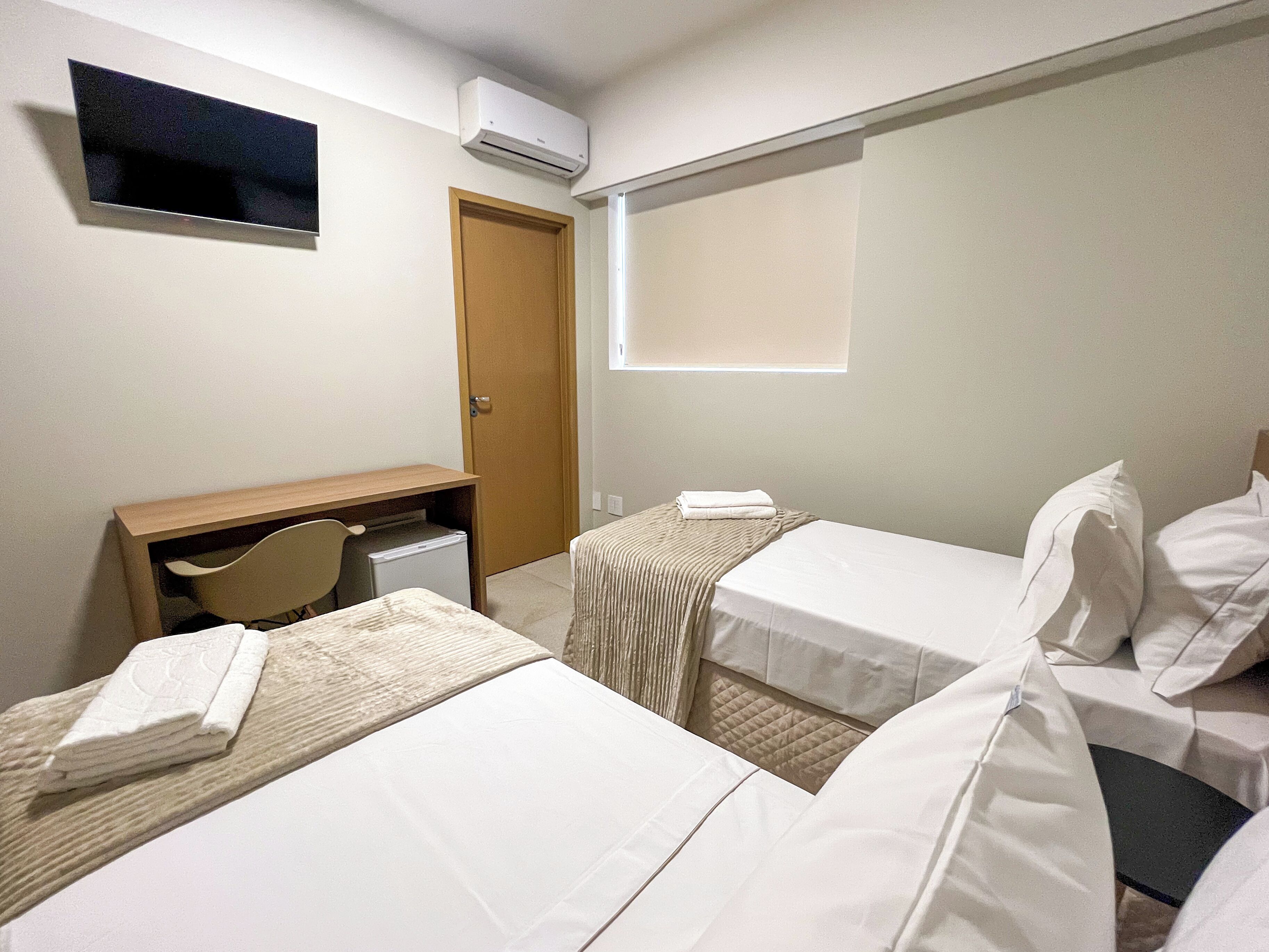 A hotel room with two single beds, a desk with a chair, a small refrigerator, and an air conditioning unit on the wall.