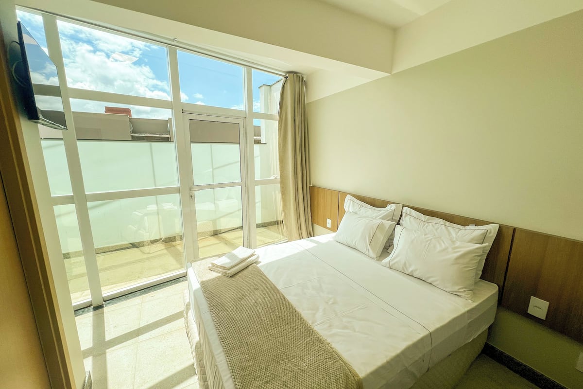 A hotel room with a large window, a bed with white linens, and a view of the sky and clouds.