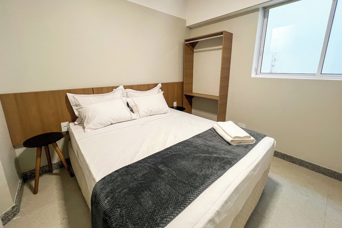 A hotel room with a bed, a small stool, a wooden headboard, a window, and a built-in shelf.