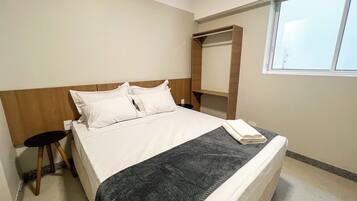 Habitación de hotel con una cama, un taburete pequeño, cabecera de madera, una ventana y una estantería empotrada.