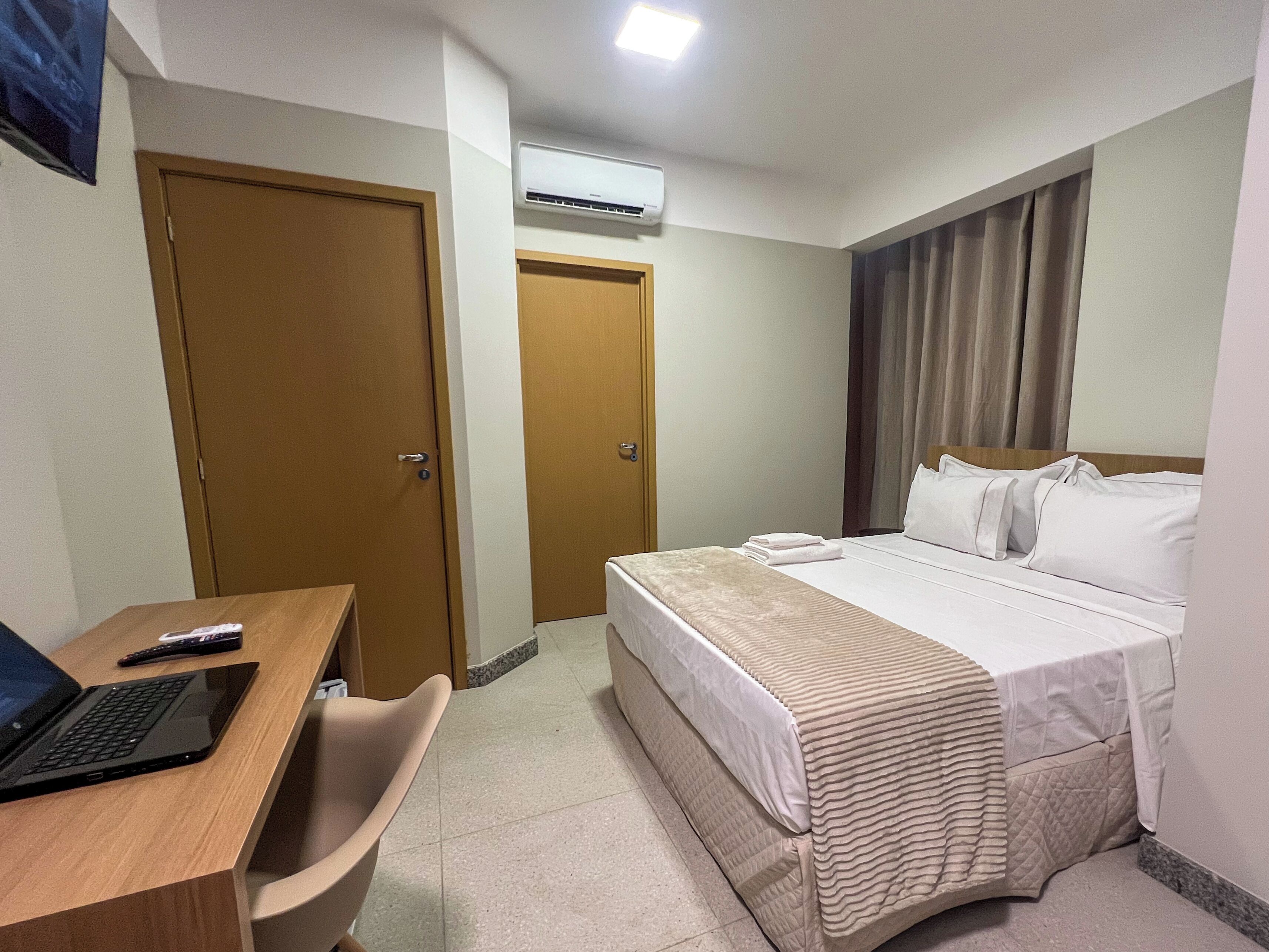 A hotel room with a bed, a desk with a laptop, a chair, and an air conditioning unit.