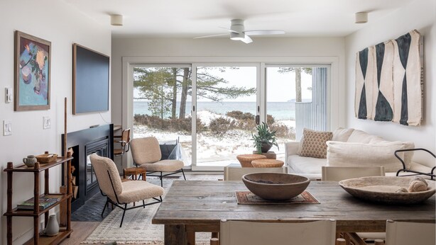 A spacious living room with a large wooden table, comfortable seating, a fireplace, and a view of the beach through large sliding glass doors.