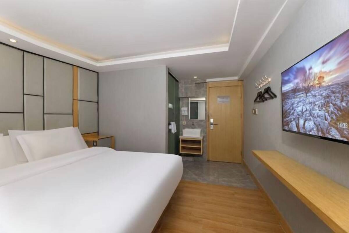 A modern hotel room with a large bed, a TV mounted on the wall, a wooden bench, and a view of the hallway.