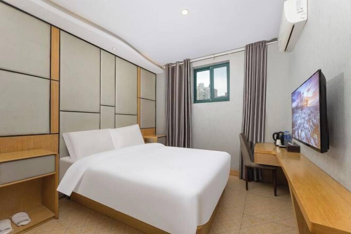 A hotel room with a bed, a desk, a chair, a TV, and an air conditioning unit.