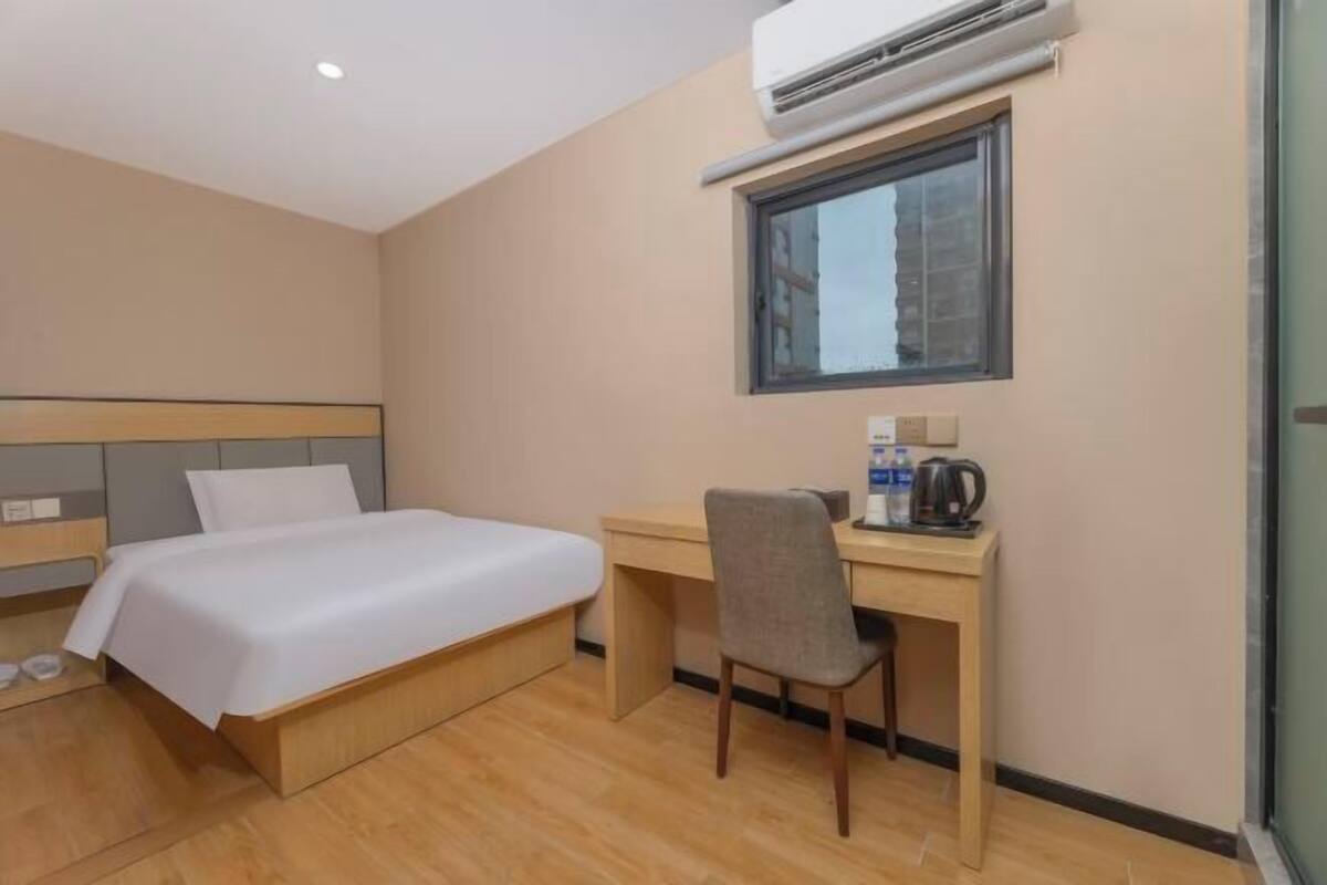 A hotel room with a bed, a desk, a chair, an air conditioning unit, and a window.