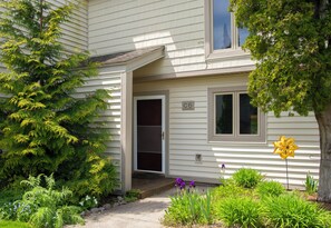 Une maison dont la porte porte le numéro « C6 », avec un porche doté de marches et un jardin où poussent diverses plantes et fleurs.