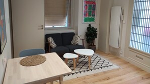 Kompakti olohuone, jossa on sohva, pieni pöytä, ruukkukasvi ja ikkuna, jossa on sälekaihtimet.
