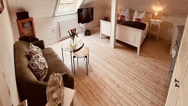 A cozy room with a sofa, a small table, a bed with a white headboard, and a television. - Vacation apartment "Still Hörn 1" in Greetsiel (approx. 30 sqm / 2 persons) (Krummhörn)
