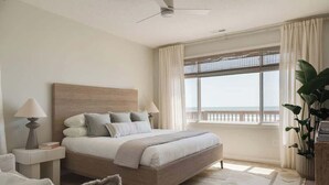 A bedroom with a bed, bedside tables, a nightstand, a chair, a potted plant, and a view of the sea through a window. - Oceanfront 7-bed home with grass backyard & grill (Folly Beach)