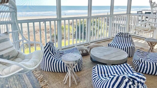 A sunlit outdoor seating area with a view of the beach, white wicker furniture, and striped cushions. - Oceanfront 7-bed home with grass backyard & grill (Folly Beach)