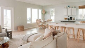A spacious living area with a large sofa, a dining table, and a kitchen area with white cabinets and a bar stool. - Oceanfront 7-bed home with grass backyard & grill (Folly Beach)