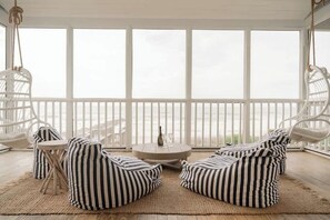 A room with a large window, white wicker chairs, a round table with a bottle, and striped cushions.