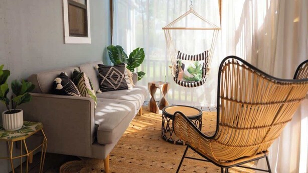 Ein gemütliches Wohnzimmer mit einer Couch, einem Hängesessel, einem kleinen runden Tisch und einem großen Fenster mit transparenten Vorhängen.