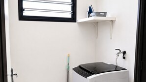 A compact laundry room with a washing machine, a drying rack, and a broom. - Roche Kerlan Apartments (Praslin Island)