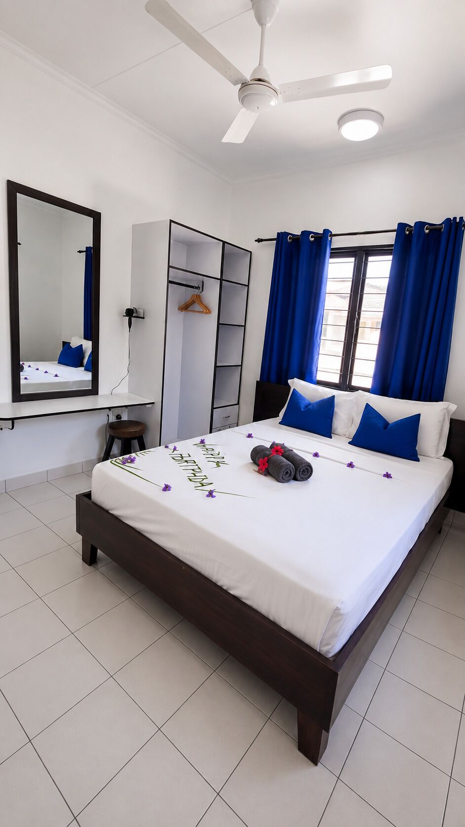 A bedroom with a bed, a wardrobe, a mirror, a ceiling fan, and a window with curtains.