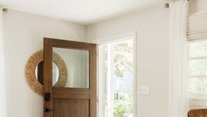 A room with a wooden door, a mirror, a wicker chair, and a view of the outside.