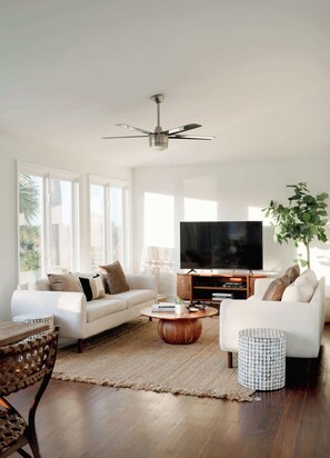 A modern living room with a white sofa, a coffee table, a TV, and a ceiling fan. - Marshfront 5BD Oasis | Pool, Yard & Steps to Beach (Folly Beach)