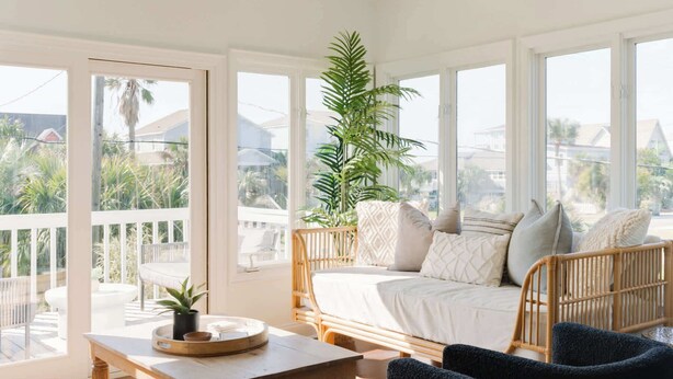 A room with a bed, a table, chairs, and large windows with a view of palm trees and houses.