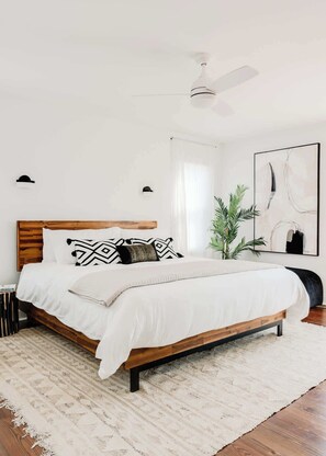 A bedroom with a bed, a ceiling fan, a window with sheer curtains, a potted plant, and a framed abstract painting. - Marshfront 5BD Oasis | Pool, Yard & Steps to Beach (Folly Beach)