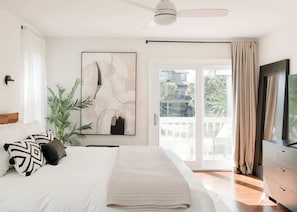 A bedroom with a bed, a ceiling fan, a large window with curtains, a plant, and a painting.