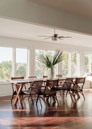 Un coin repas avec une table en bois et des chaises en osier, de grandes fenêtres et un ventilateur de plafond.