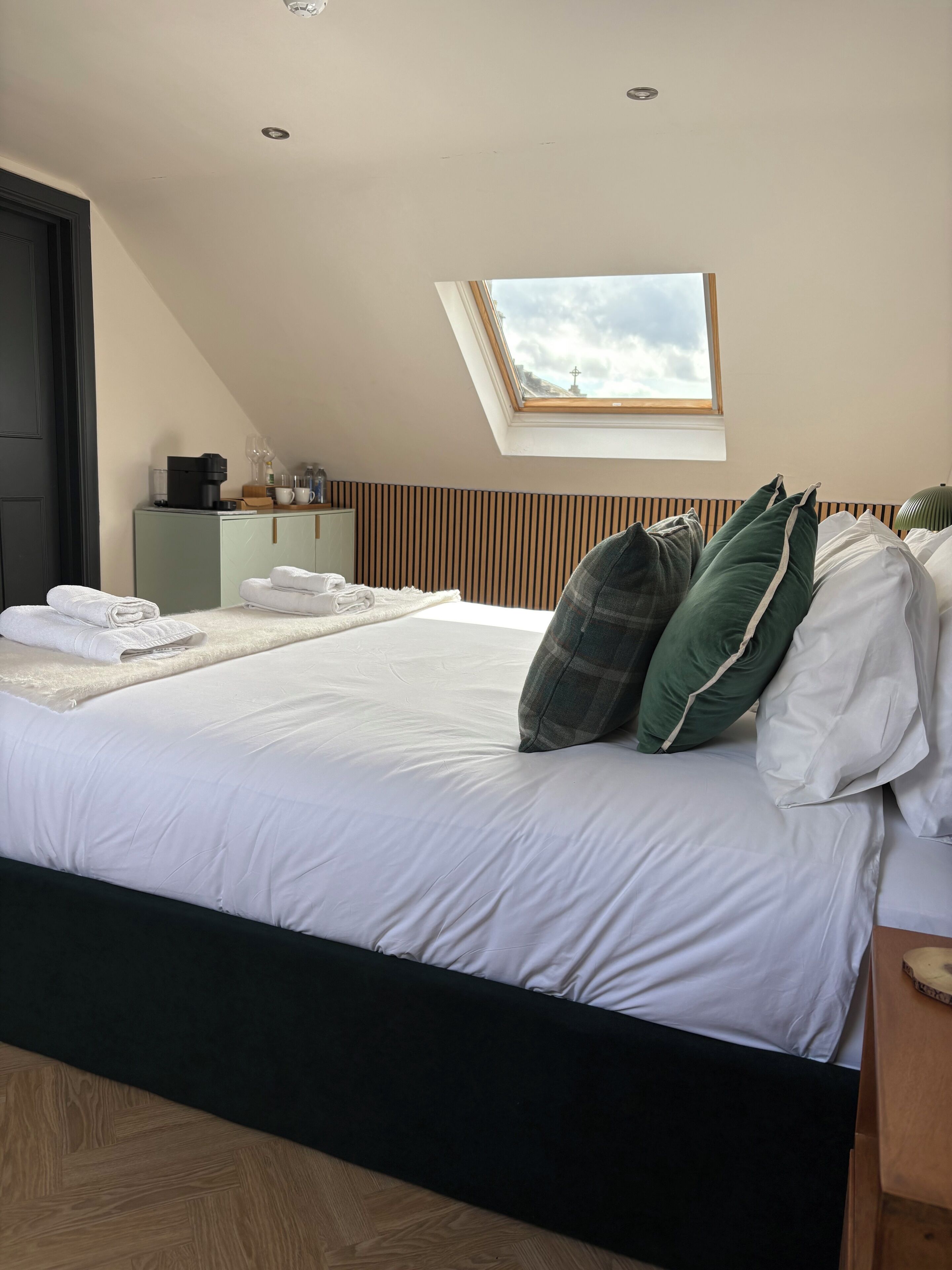 A bedroom with a bed, pillows, a desk with a lamp, a window, and a door.
