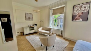 A spacious living room with a white sofa, a white armchair, a wooden herringbone floor, a white cabinet, and a large window with a view of greenery. - ApartNest Auchterarder (Auchterarder)