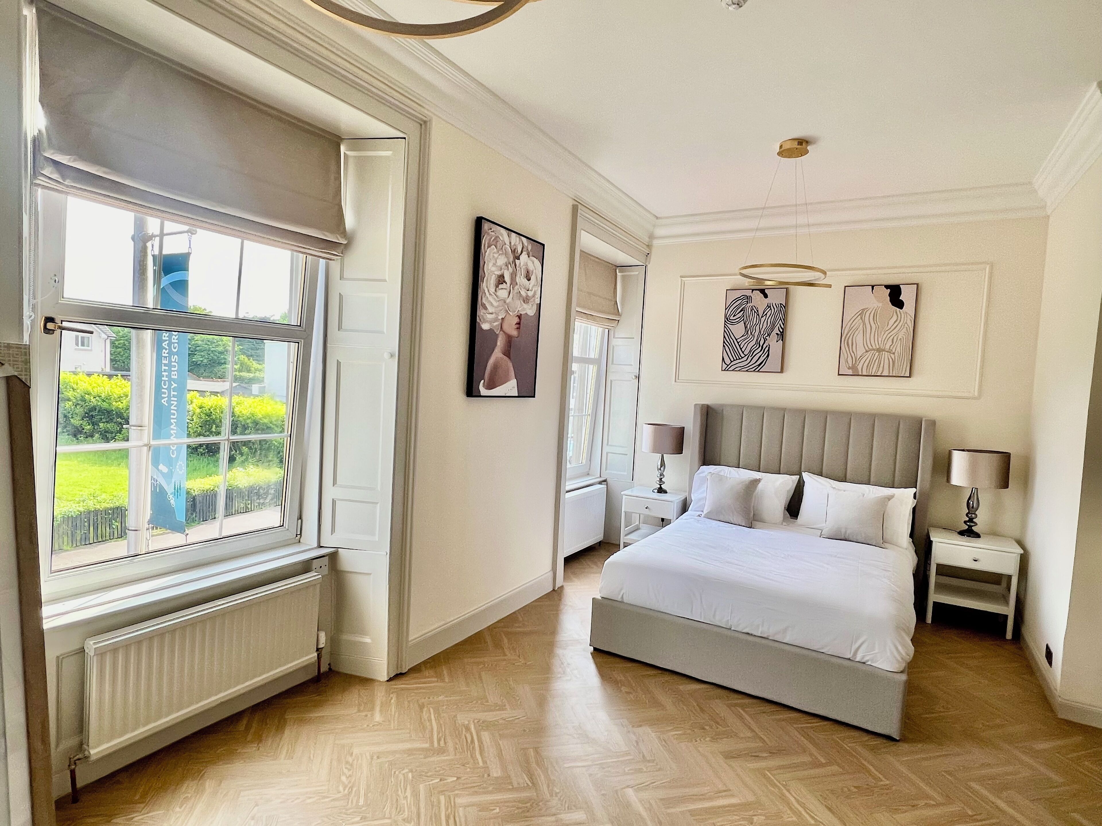 A bedroom with a large bed, two bedside tables, a radiator, and a window with a view of greenery.