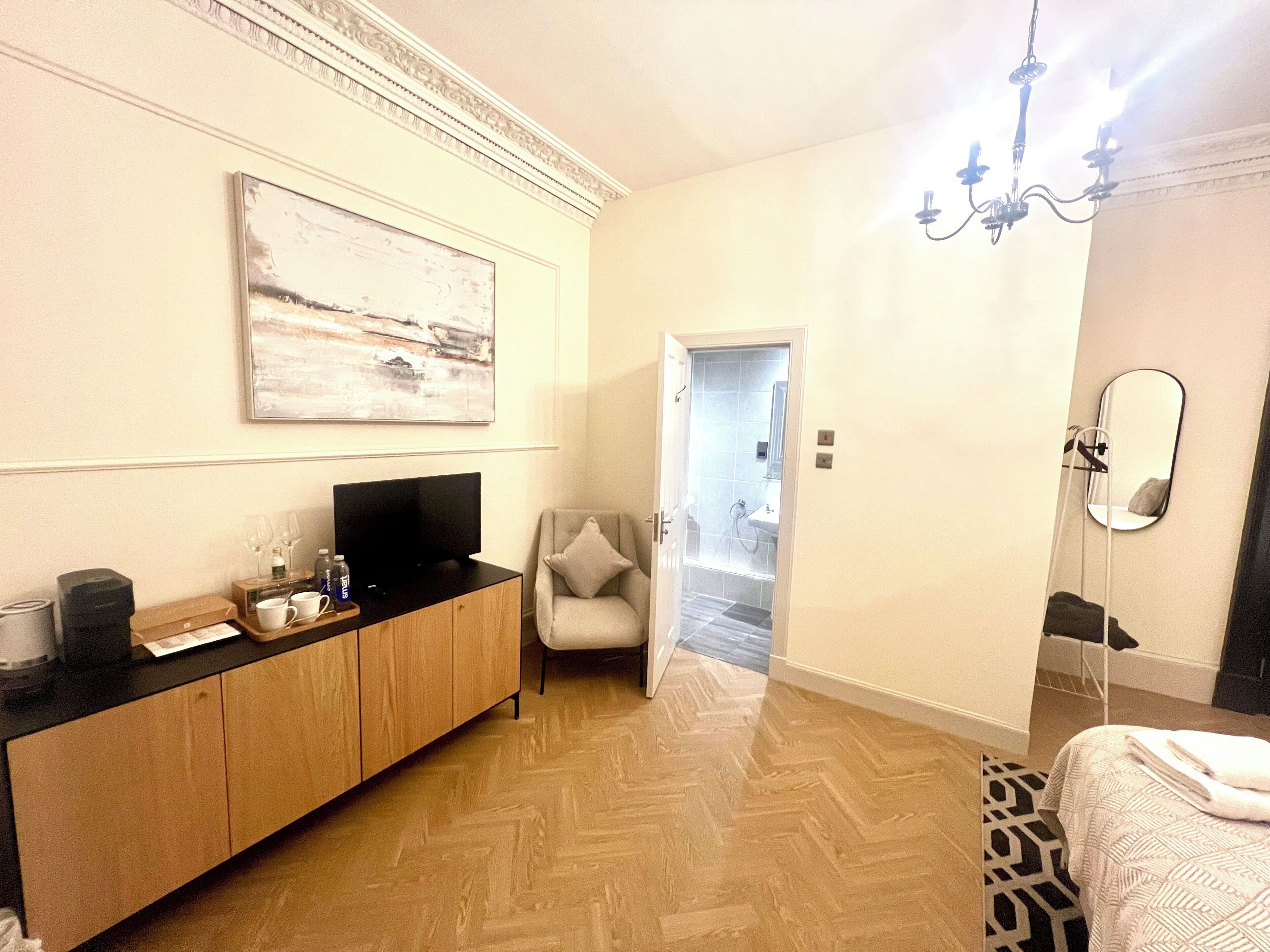 A hotel room with a wooden herringbone floor, a black TV on a wooden cabinet, a white chair, a door leading to a bathroom, and a mirror on the wall.