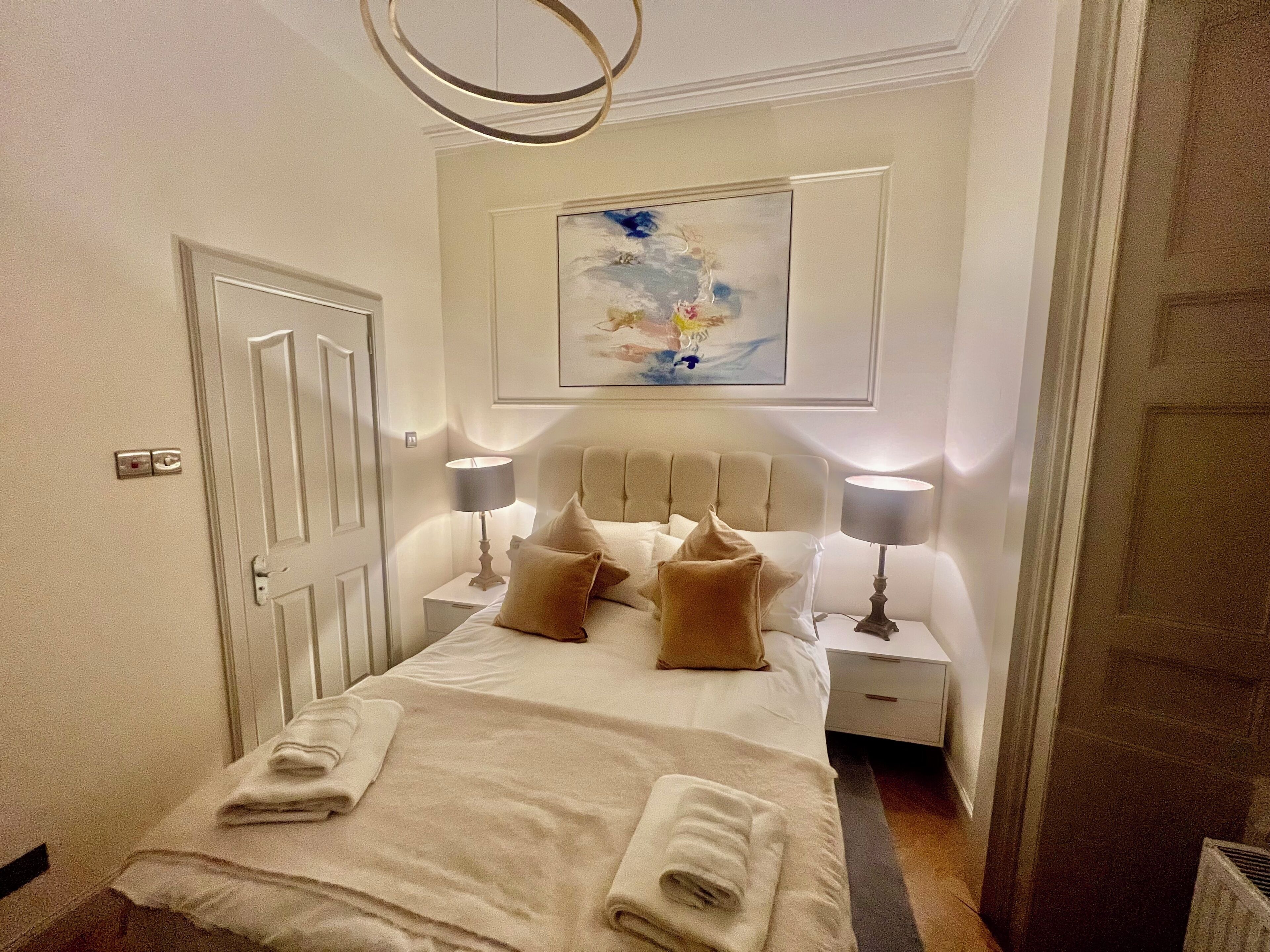 A neatly made bed with pillows and a folded towel, a bedside lamp, a nightstand, a door, and a painting on the wall.
