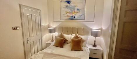 A neatly made bed with pillows and a folded towel, a bedside lamp, a nightstand, a door, and a painting on the wall.