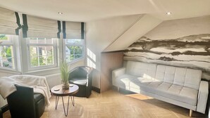 A modern loft bedroom with a large window, a grey sofa, a black chair, and a wooden herringbone floor. - ApartNest Auchterarder (Auchterarder)