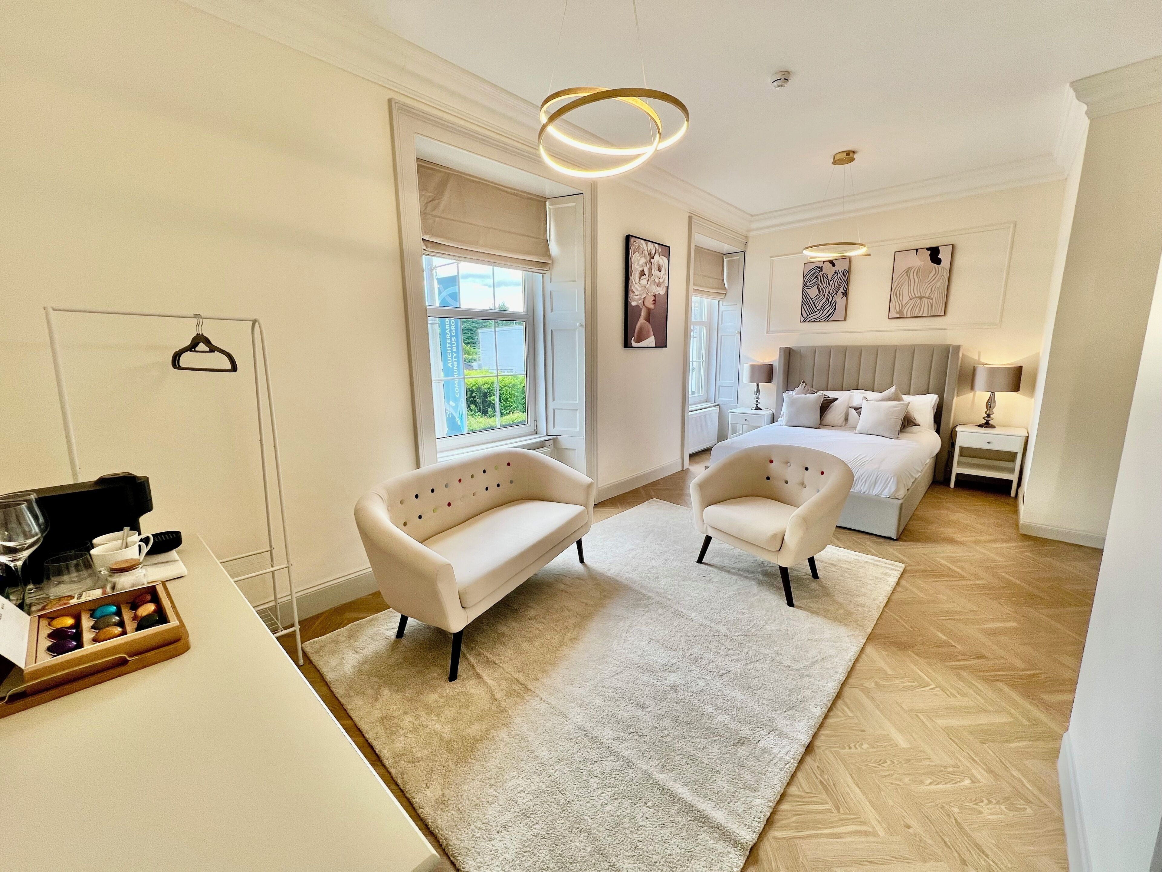 A modern hotel room with a bed, two armchairs, a desk with a coffee maker, and a window with a view of greenery.