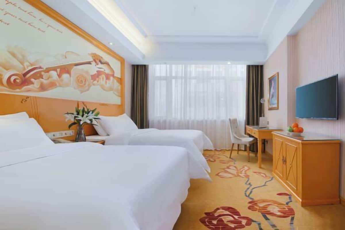 A hotel room with two beds, a desk, a TV, and a large floral artwork on the wall.