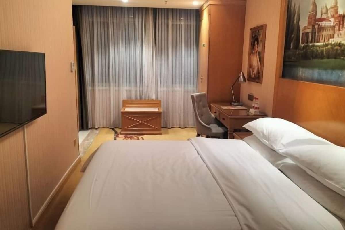 A hotel room with a bed, a desk with a lamp, a chair, a small table, and a painting on the wall.