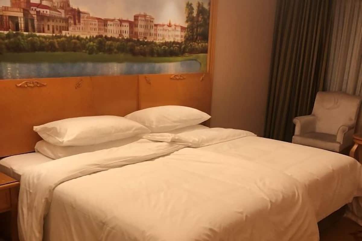 A hotel room with a bed, a nightstand, a chair, and a painting of a building and lake on the wall.