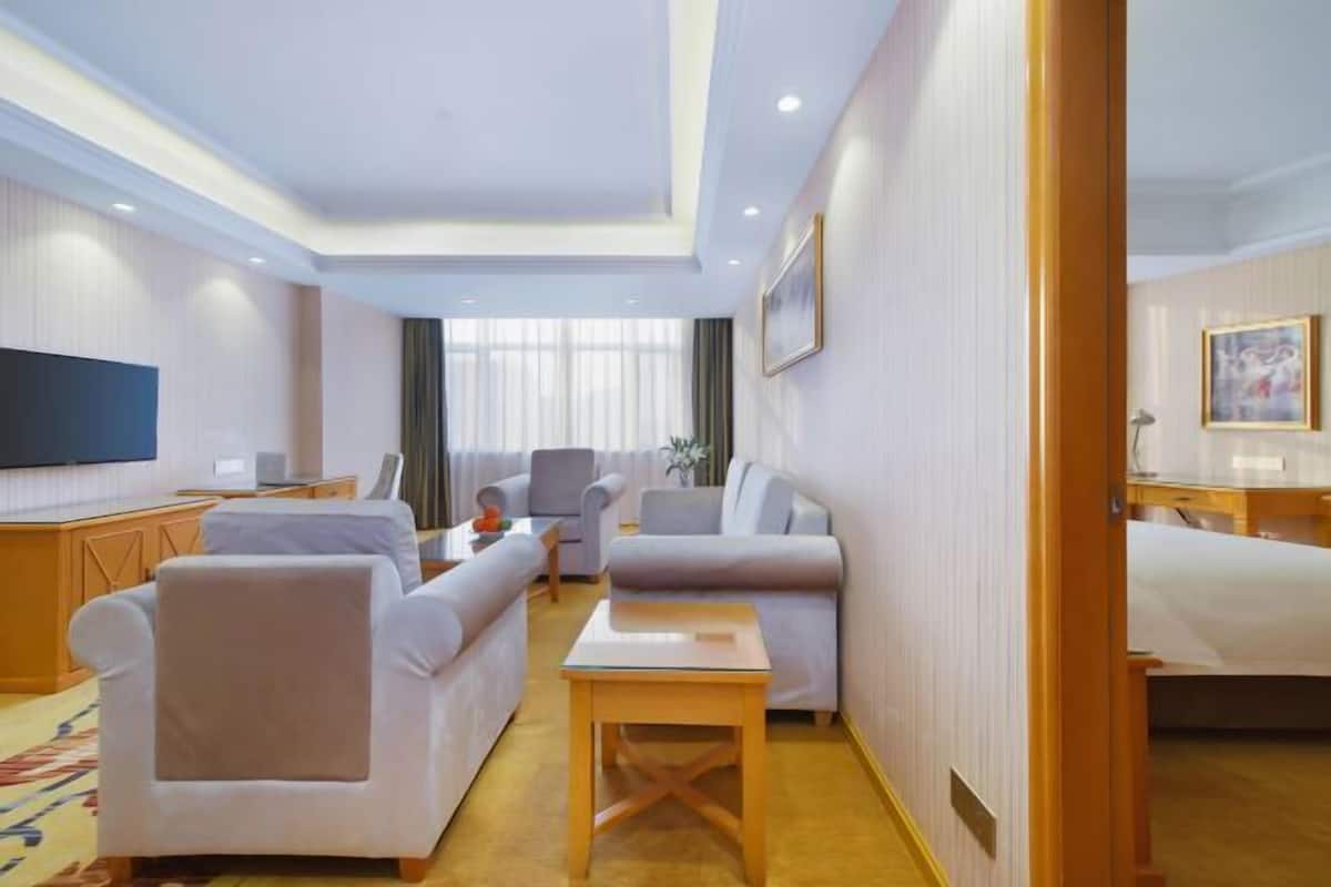 A hotel room with a large bed, a sofa, a chair, a small table, a TV, and a window with curtains.
