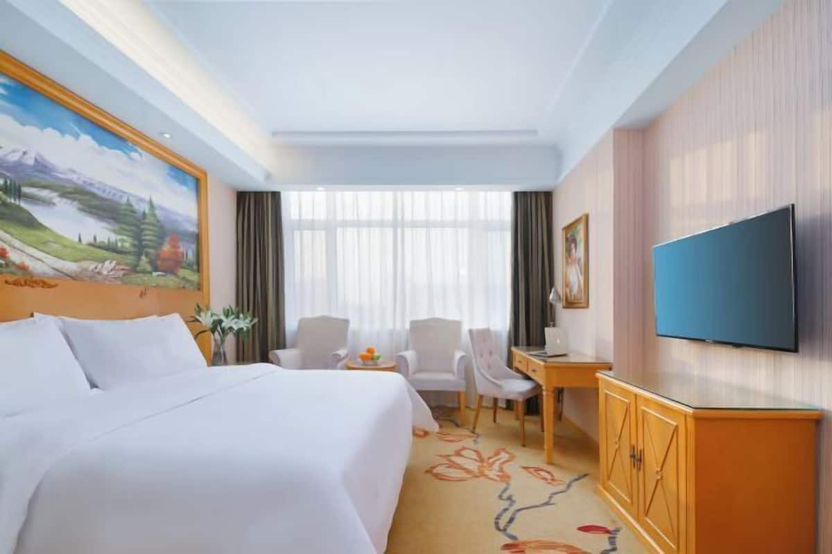 A hotel room with a bed, a desk, a chair, a television, and a painting on the wall.