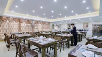 A dining area with multiple tables and chairs, a person serving, and a kitchen in the background.