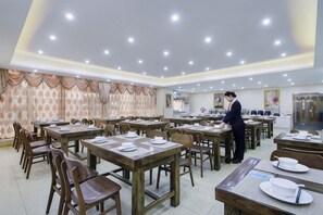 Une salle à manger avec plusieurs tables et chaises, une personne qui sert les clients, et une cuisine en arrière-plan.