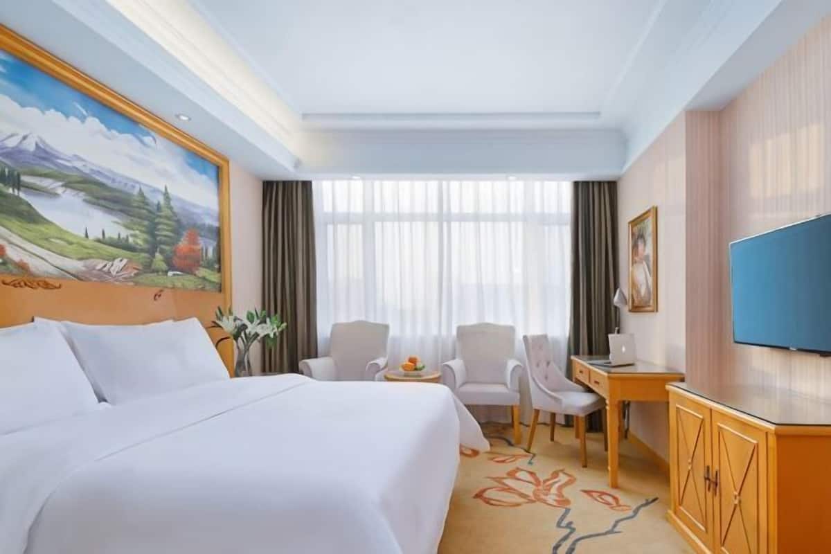 A hotel room with a large bed, a desk, two chairs, a television, and a painting on the wall.